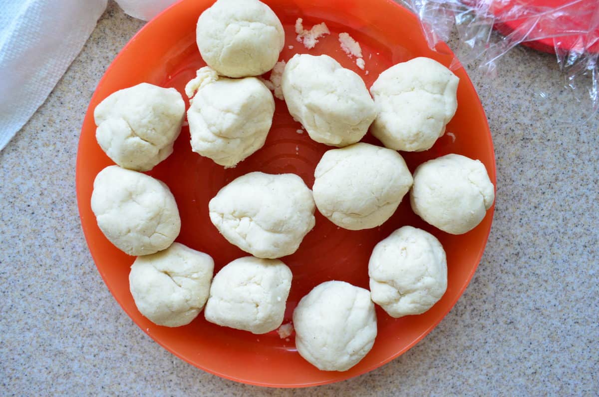 13 small balls of dough resting on an orange plate with plastic wrap next to plate.