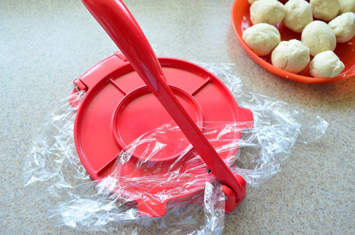 Red tortilla press closed, with plate of dough balls on the side. 