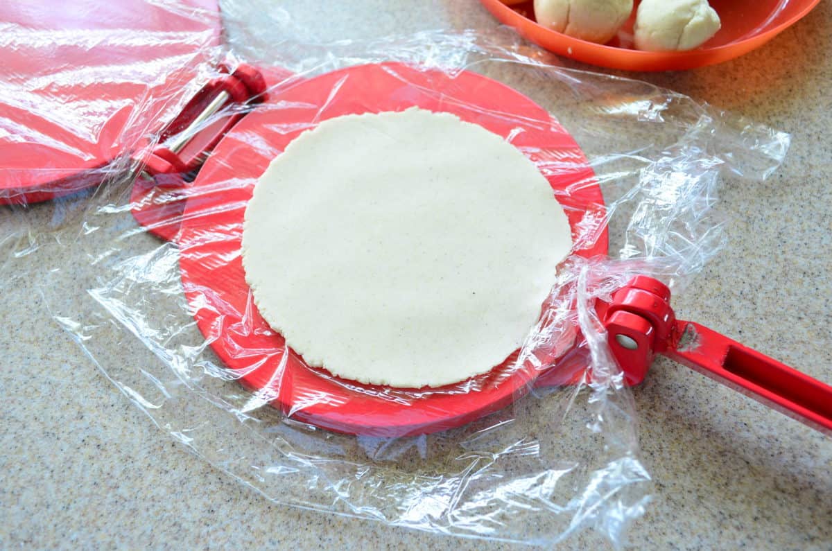 Pressed dough on plastic wrap sitting on a red tortilla press.