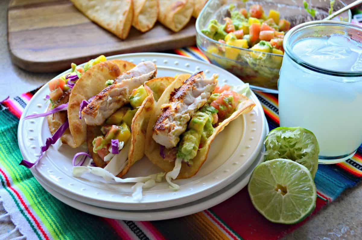 3 plated Mahi Mahi Fish Tacos with Mango Avocado Salsa next to lime and a yellow iced beverage.