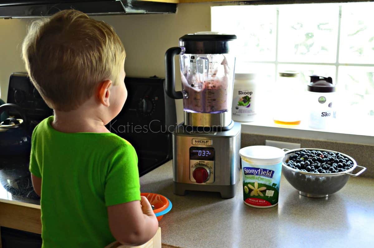 Toddler boy watching a smoothie being made in a blender with blueberries and yogurt on the counter.
