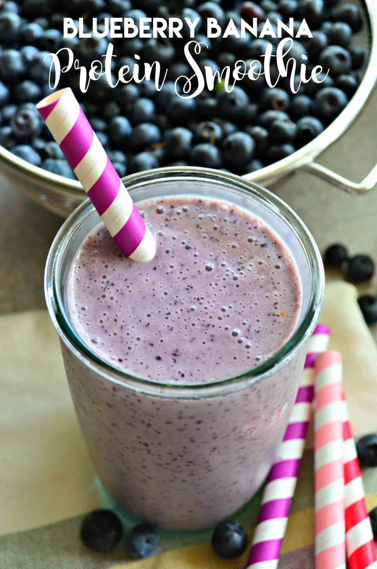 Close up of a large glass filled with a blueberry smoothie with a striped straw and fresh blueberries in background. 