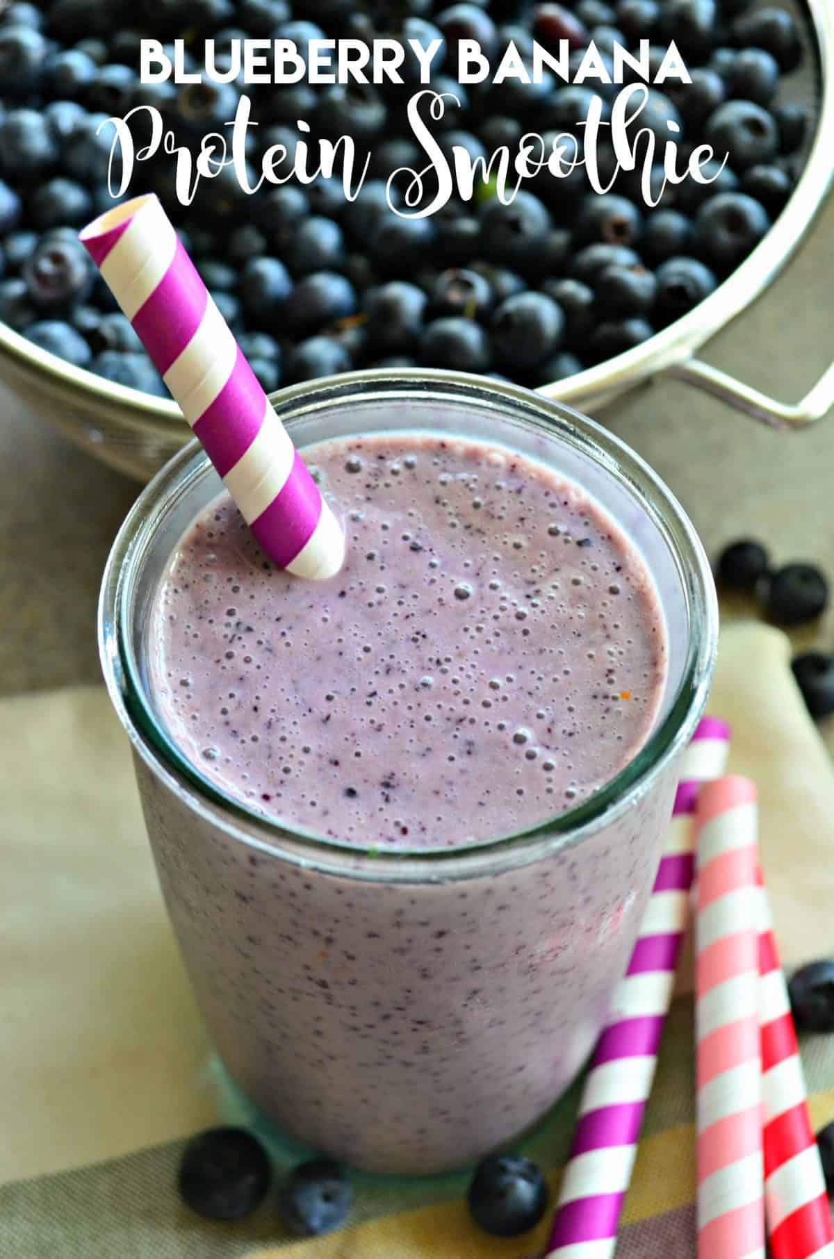 Close up of a large glass filled with a blueberry smoothie with a striped straw and fresh blueberries in background.