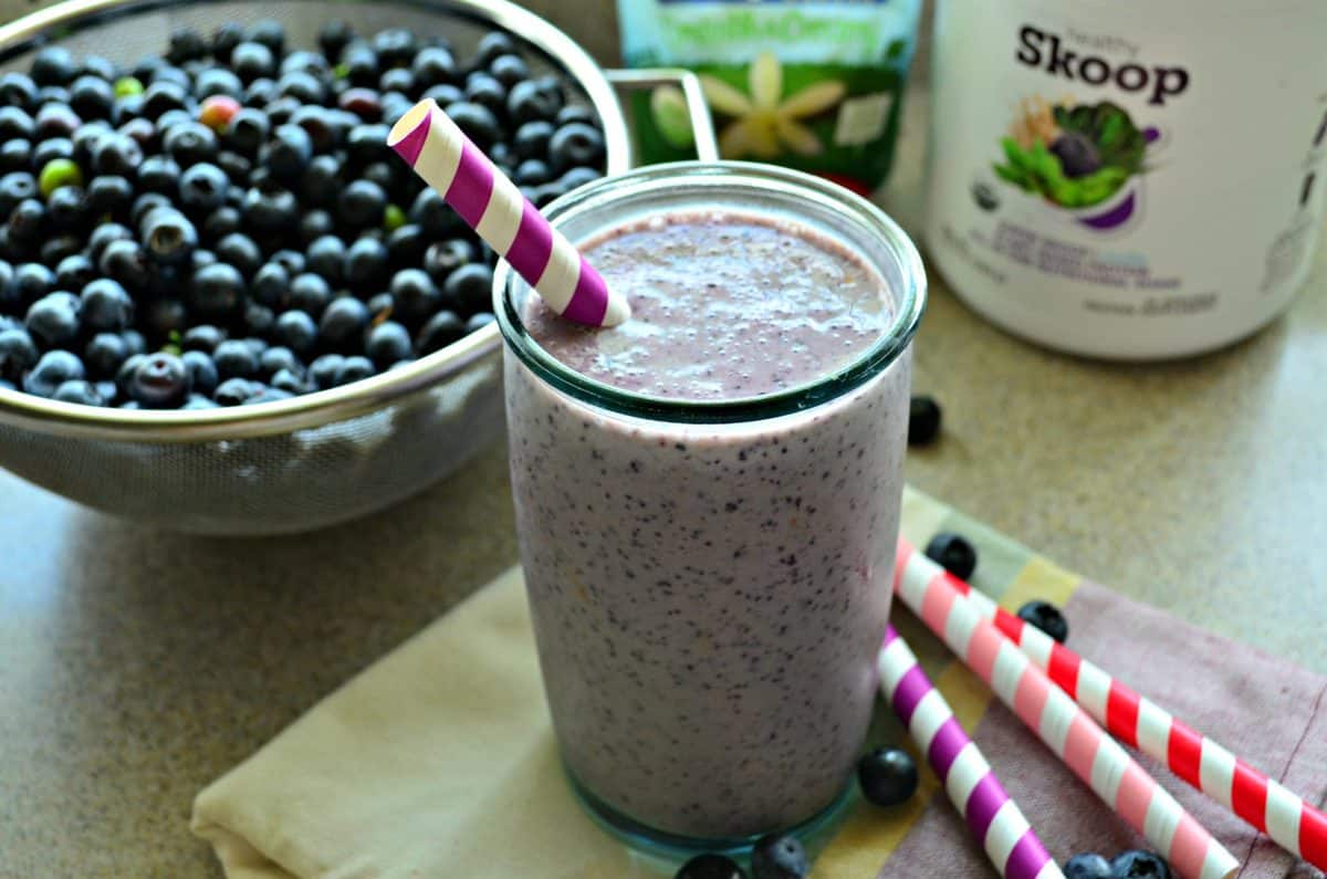 Large glass filled with a Blueberry Banana Protein Smoothie with a colander of blueberries in the background.