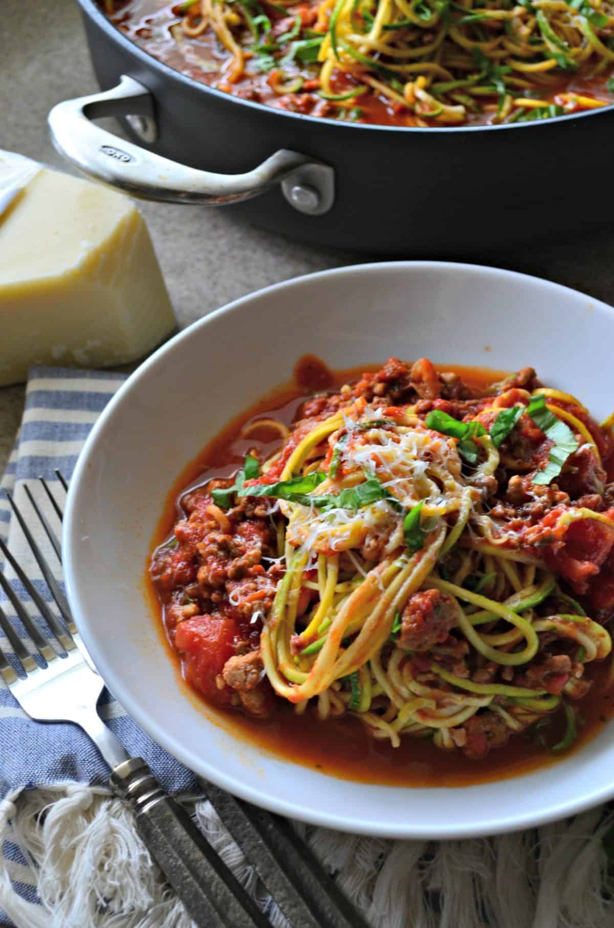 White bowl filled with zucchini noodles and meat sauce with skillet in background.