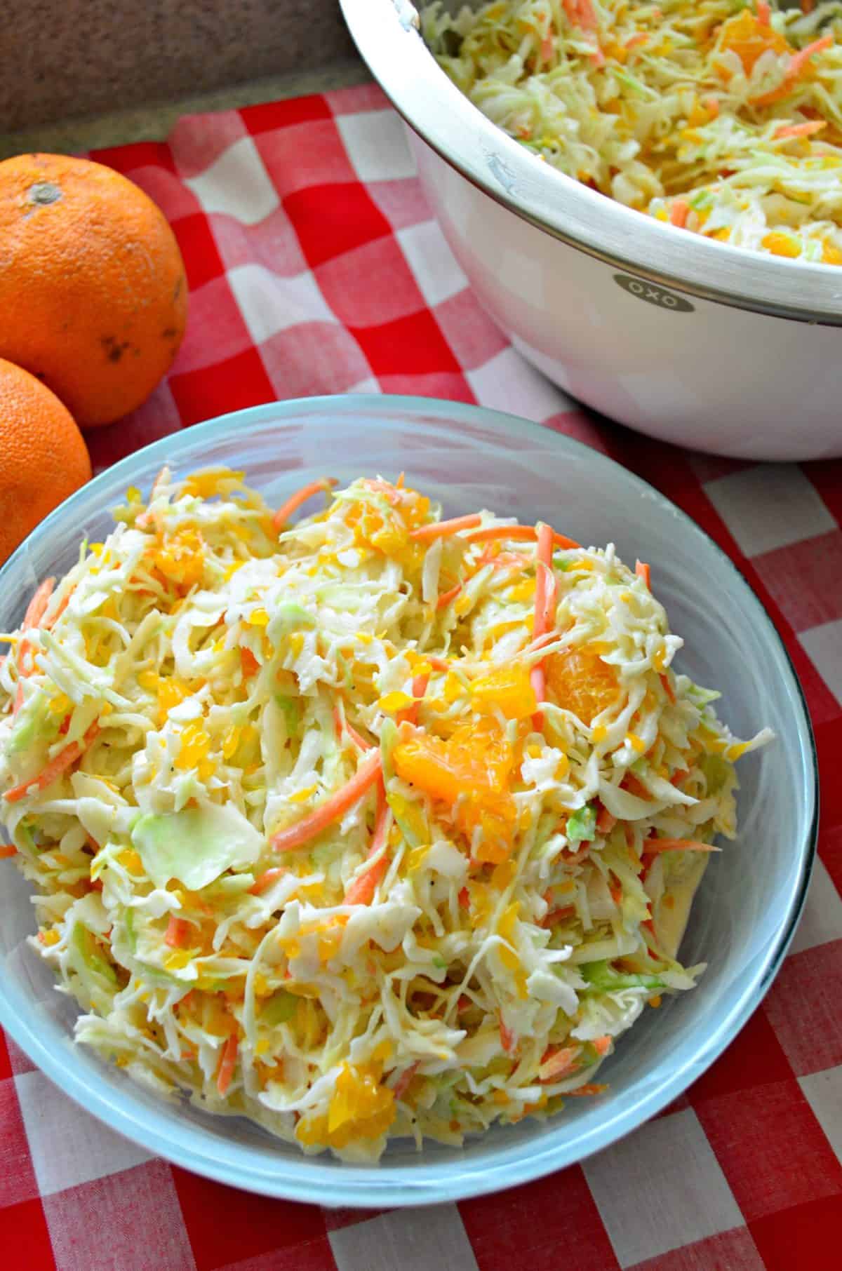 Overhead view of coleslaw with oranges on a red and white checkered cloth. 