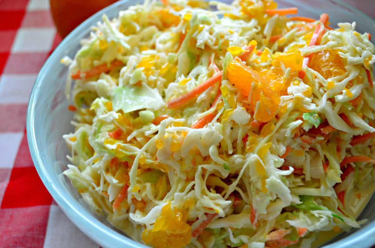 Side view of a blue bowl with coleslaw with oranges in it on a red and white checkered cloth.