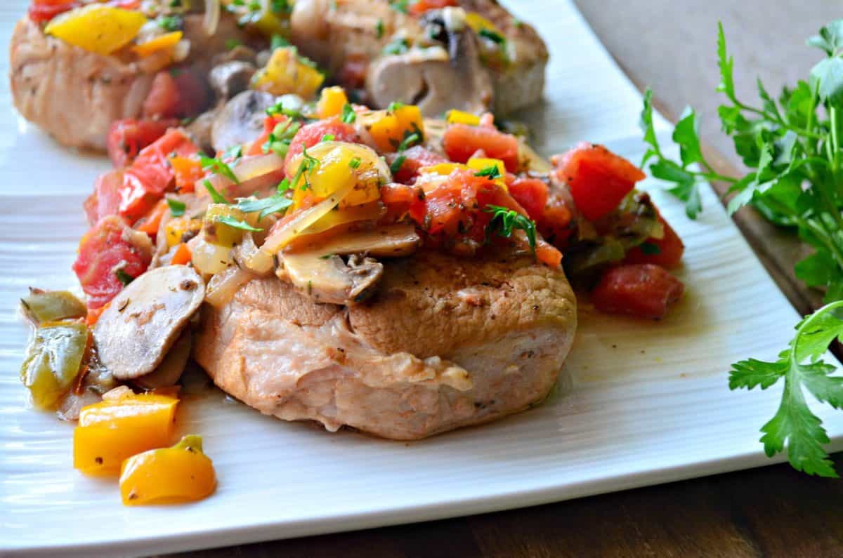 plated pork chops covered in herbs, tomatoes, mushrooms, and parsley on white plate.