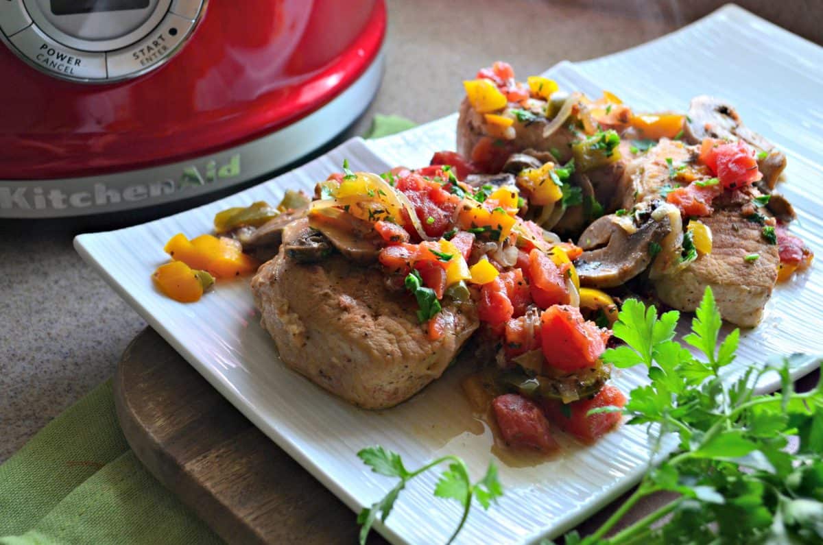 top view of pork chops covered in herbs, tomatoes, mushrooms, and parsley on white plate in front of red slow cooker.