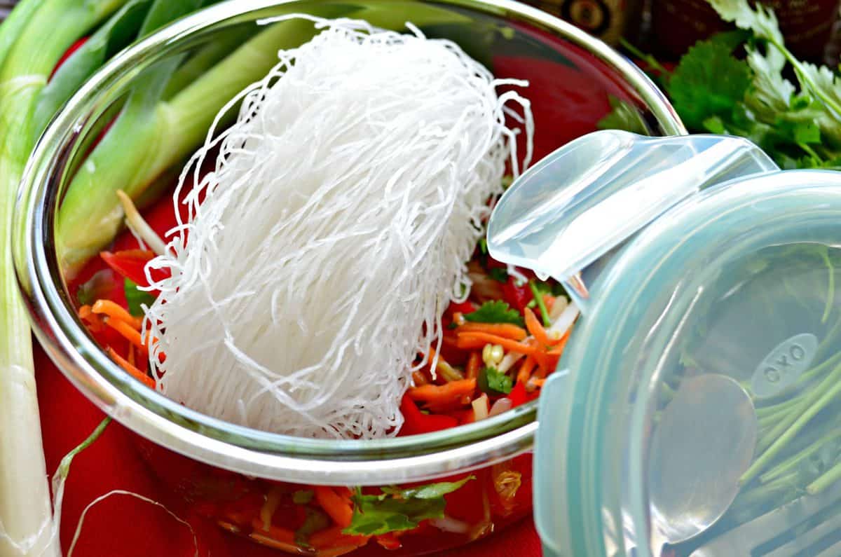 glass bowl of uncooked rice noodles with sliced peppers and cilantro.
