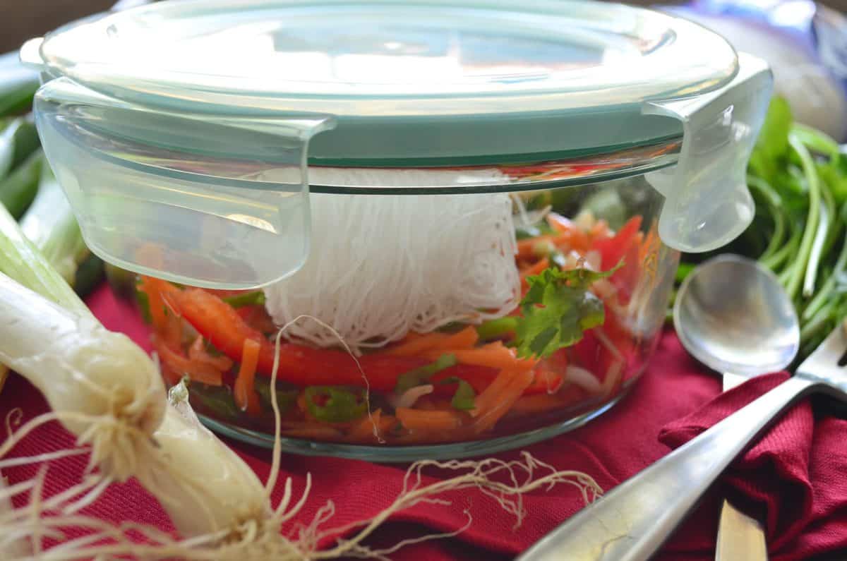 side view of closed glass container filled with uncooked sliced peppers, rice noodles, cilantro, and bean sprouts.
