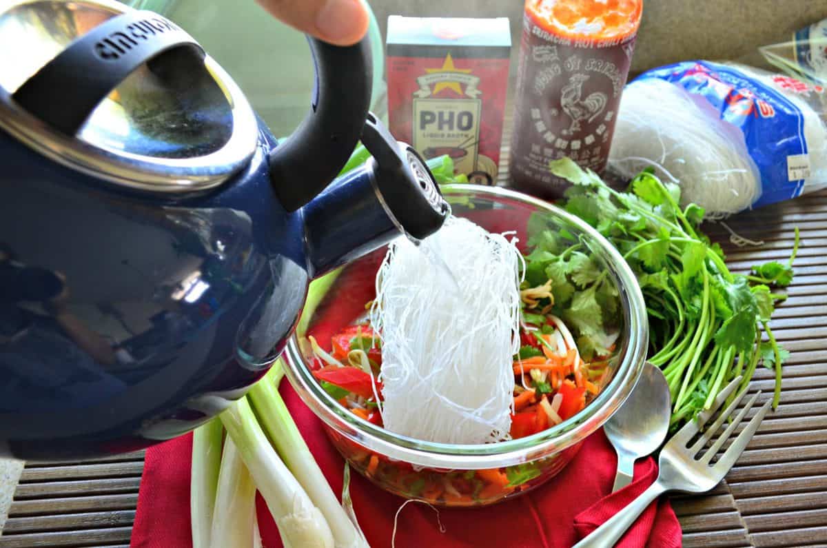 top view of kettle held over glass container filled with uncooked sliced peppers, rice noodles, cilantro, and bean sprouts.