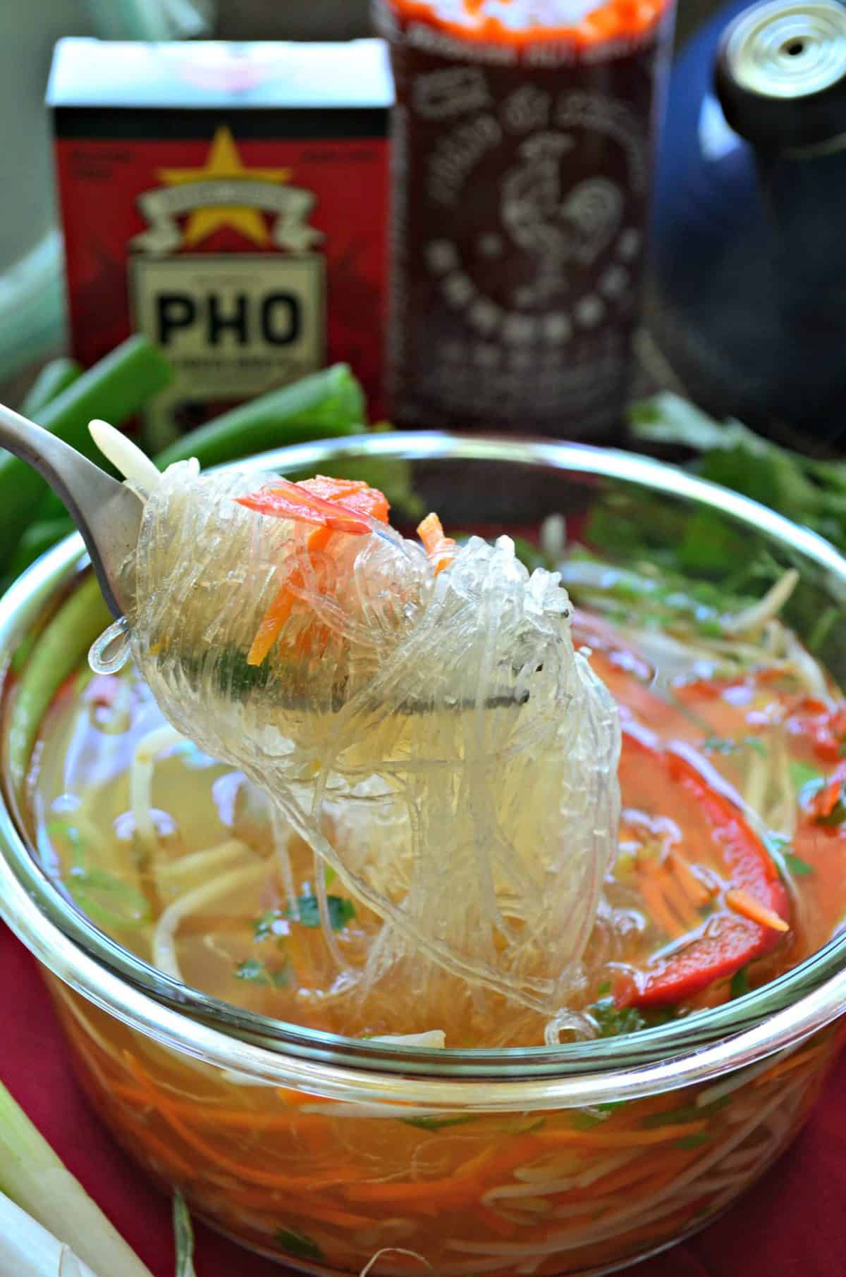 closeup fork full of rice noodles over bowl of pho filled with broth and veggies.