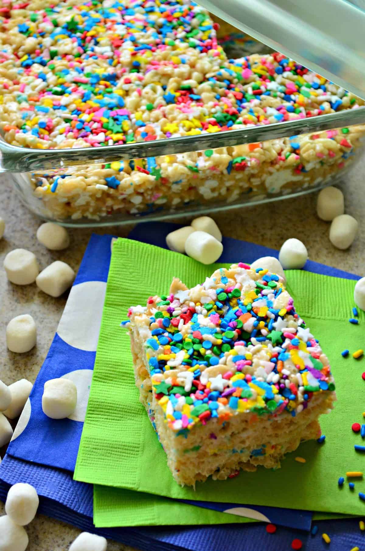 colorful rice crispy square on green napkin in front of glass casserole dish of remaining uncut rice crispy treat.