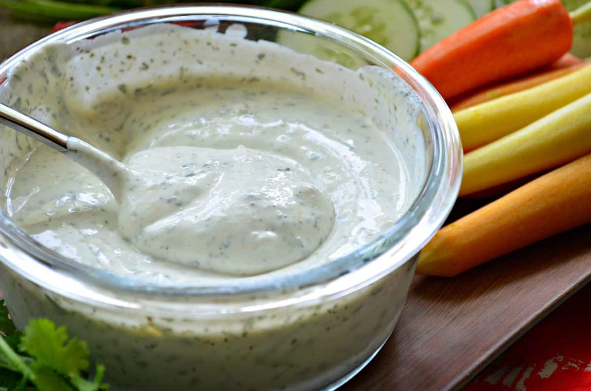 closeup of Greek Yogurt Ranch Dip in glass bowl with metal spoon, veggies in background.