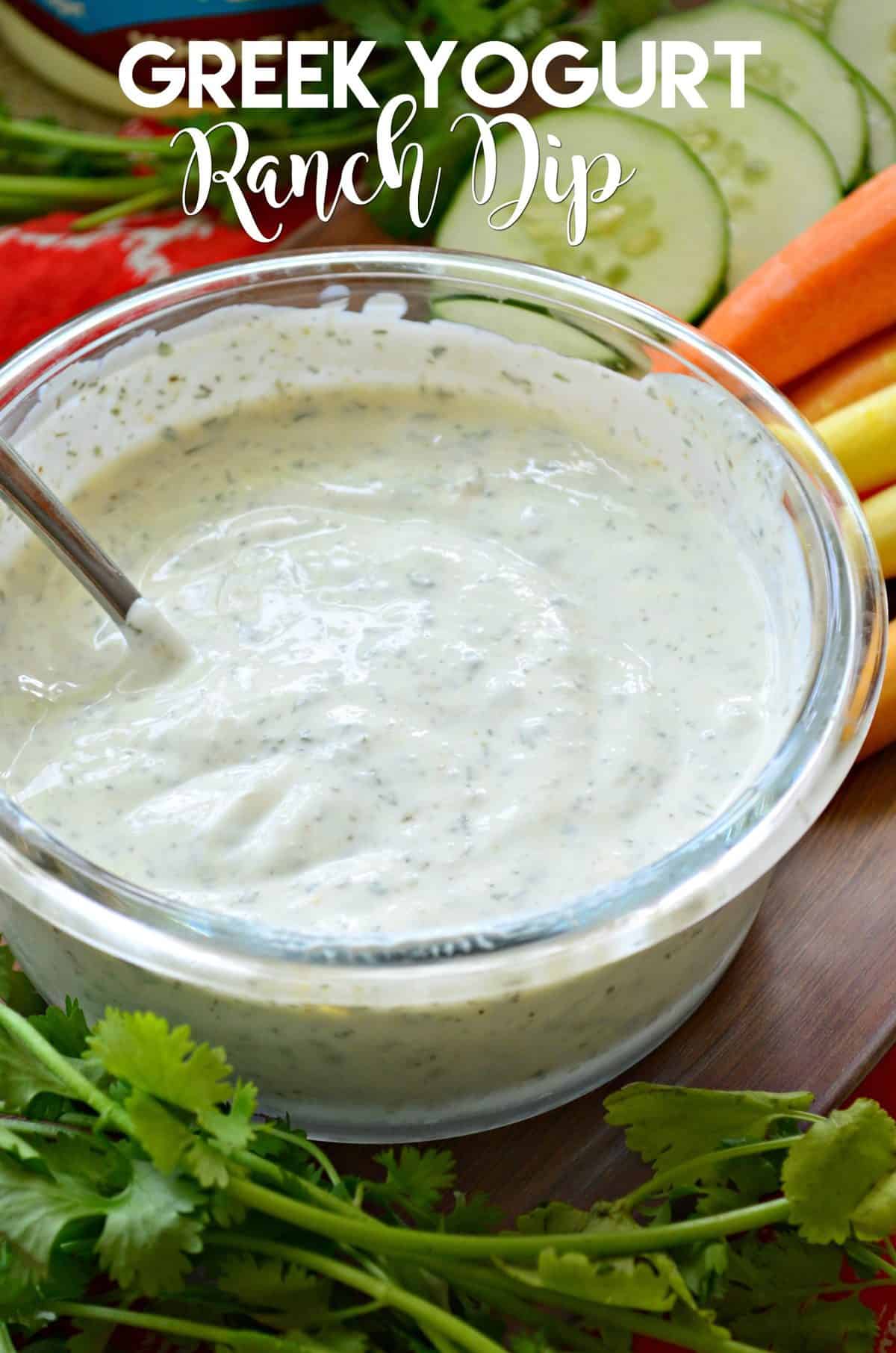 closeup of Greek Yogurt Ranch Dip in glass bowl with metal spoon, veggies in background, and title text.