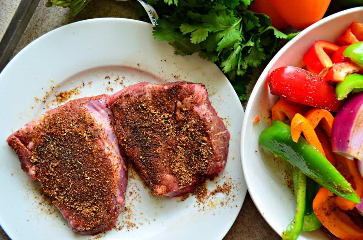 top view of 2 plated slices of uncooked steak dusted with seasonings next to bowl of uncooked veggies.