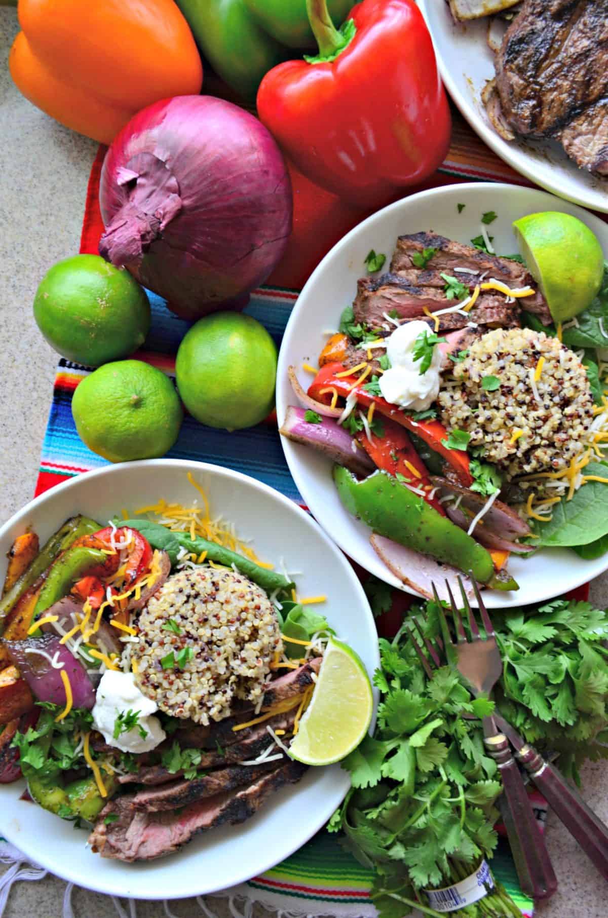 top view 2 white bowls with quinoa, grilled onions and peppers, steak, cilantro, and sour cream next to fresh veggies.