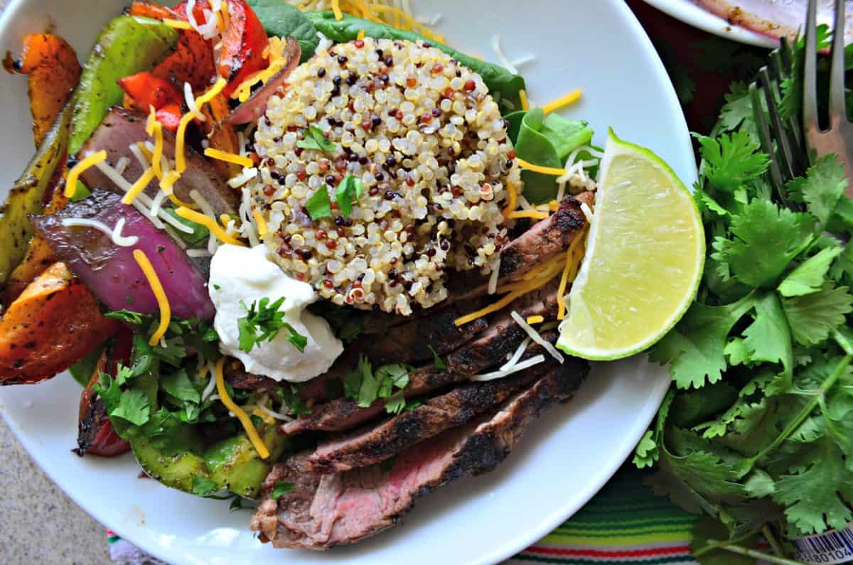 top view of white bowl of quinoa, steak, grilled veggies, sour cream, cilantro, and lime.