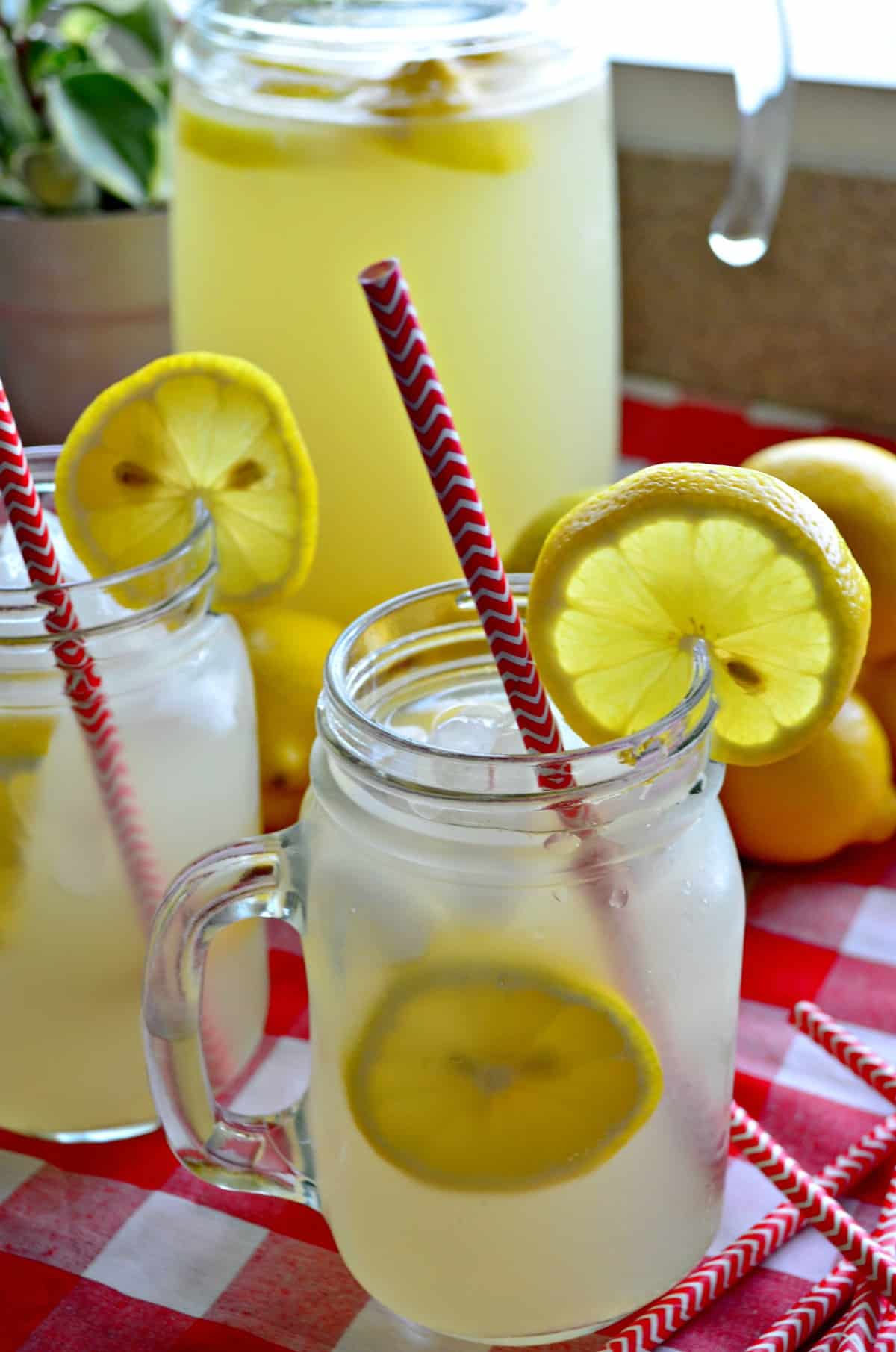 Closeup side view of lemonade in two mason jars with fresh lemon wheels and red straws and pitcher in background.