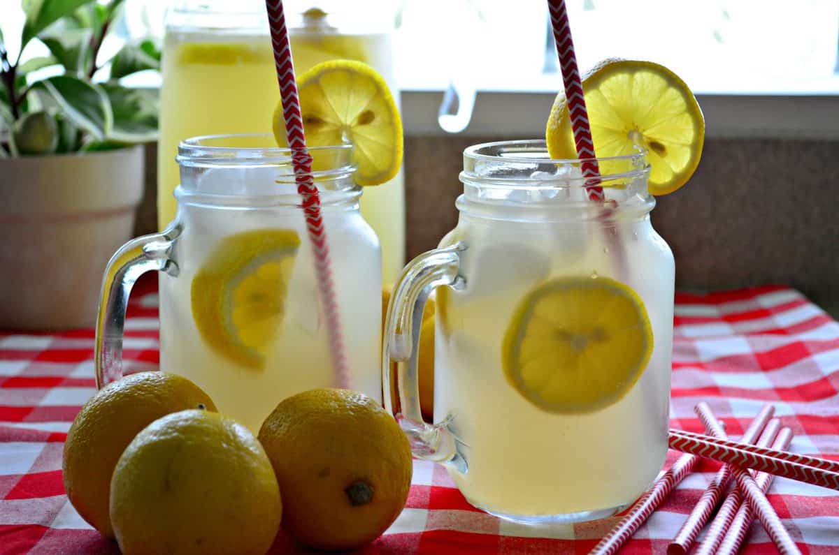 side view of lemonade in two mason jars with fresh lemon wheels and red straws on checkered tablecloth.