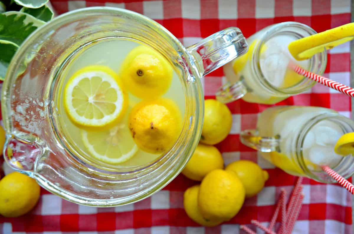 Top view pitcher of lemonade with fresh lemon wheels next to two jars of lemonade on tablecloth.