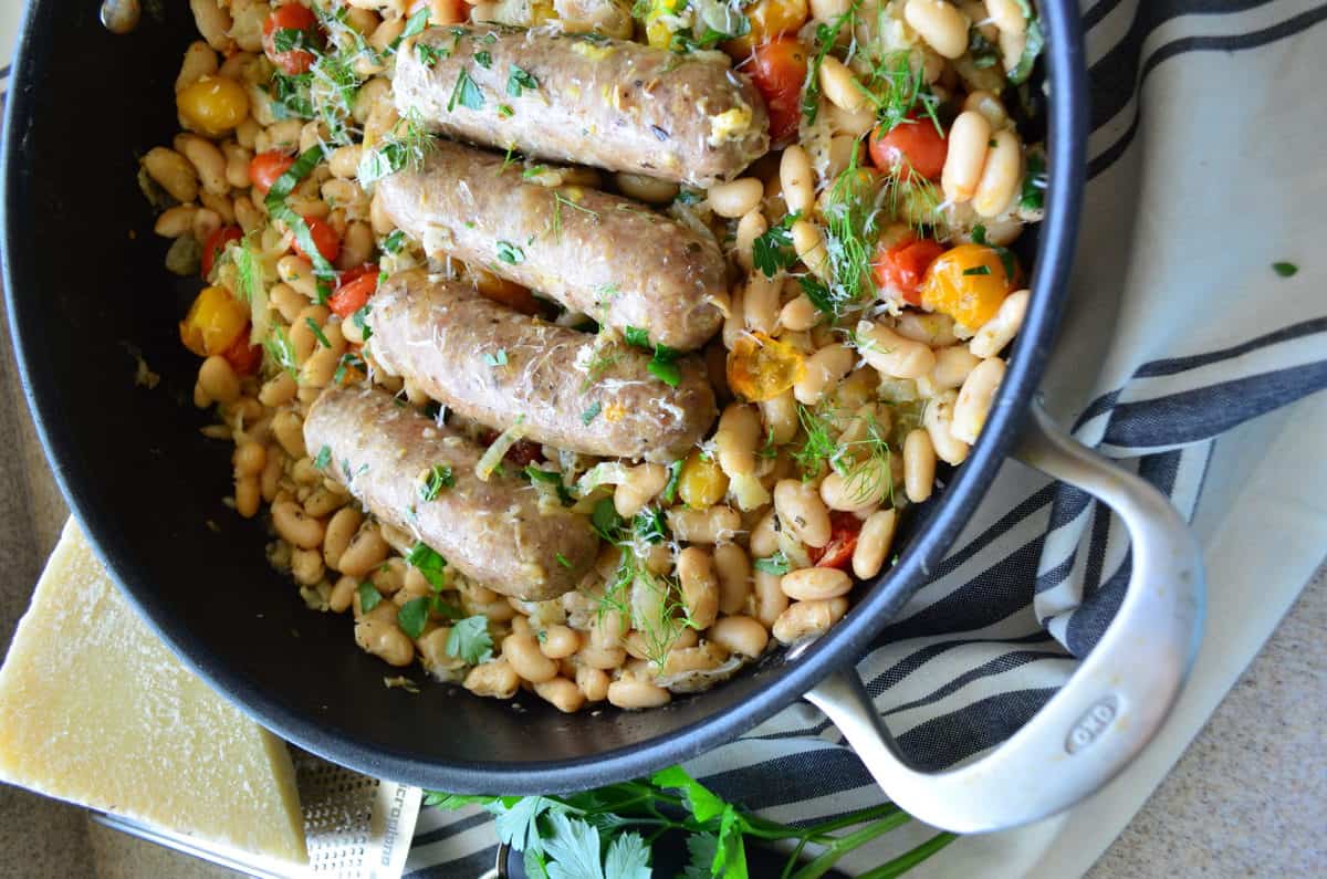 sideways top view of 5 sausages in skillet over beans with basil and cheese next to parsley on tablecloth.