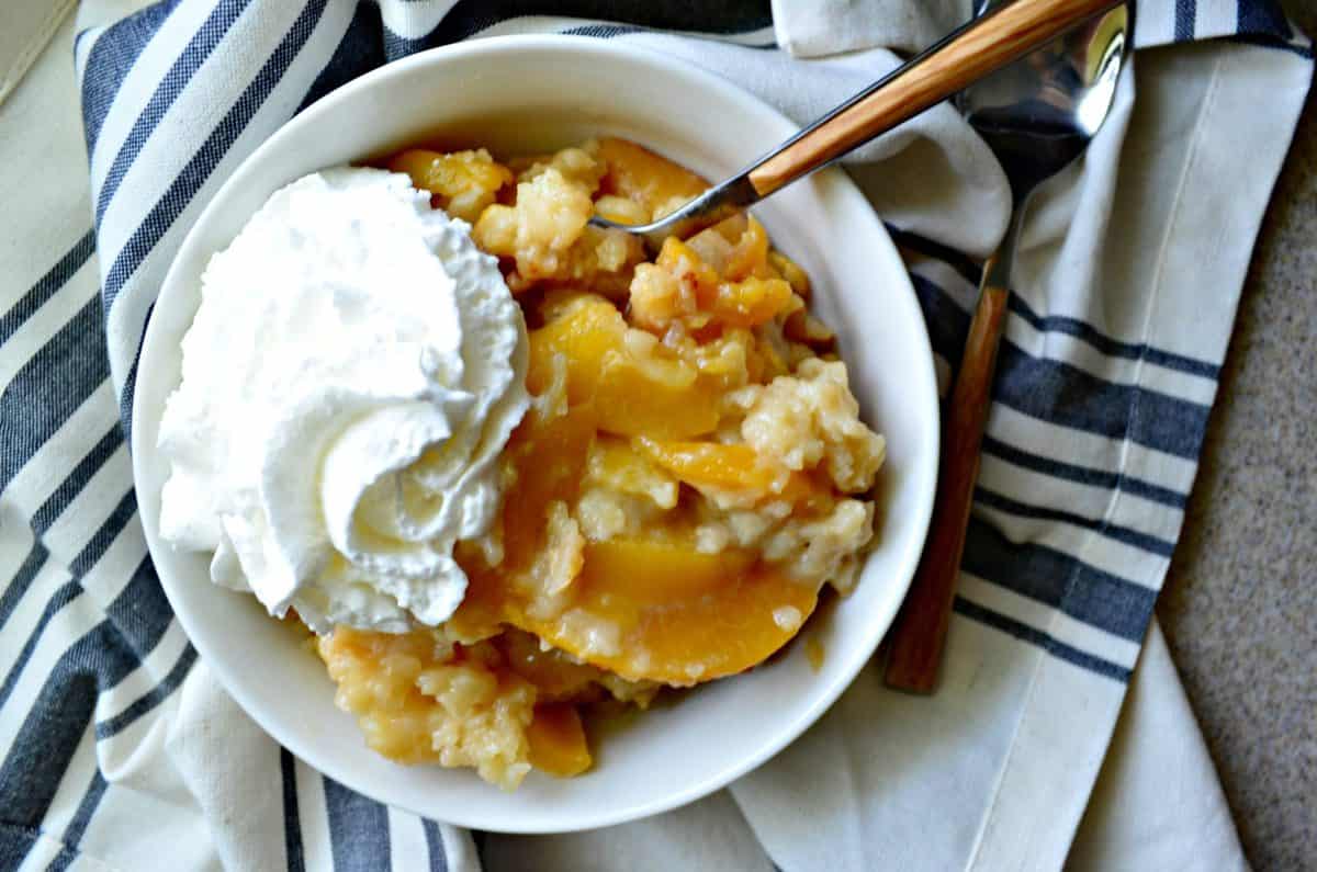 Top view of white dish filled with peach slices, whipped cream, and cobbler with title text.