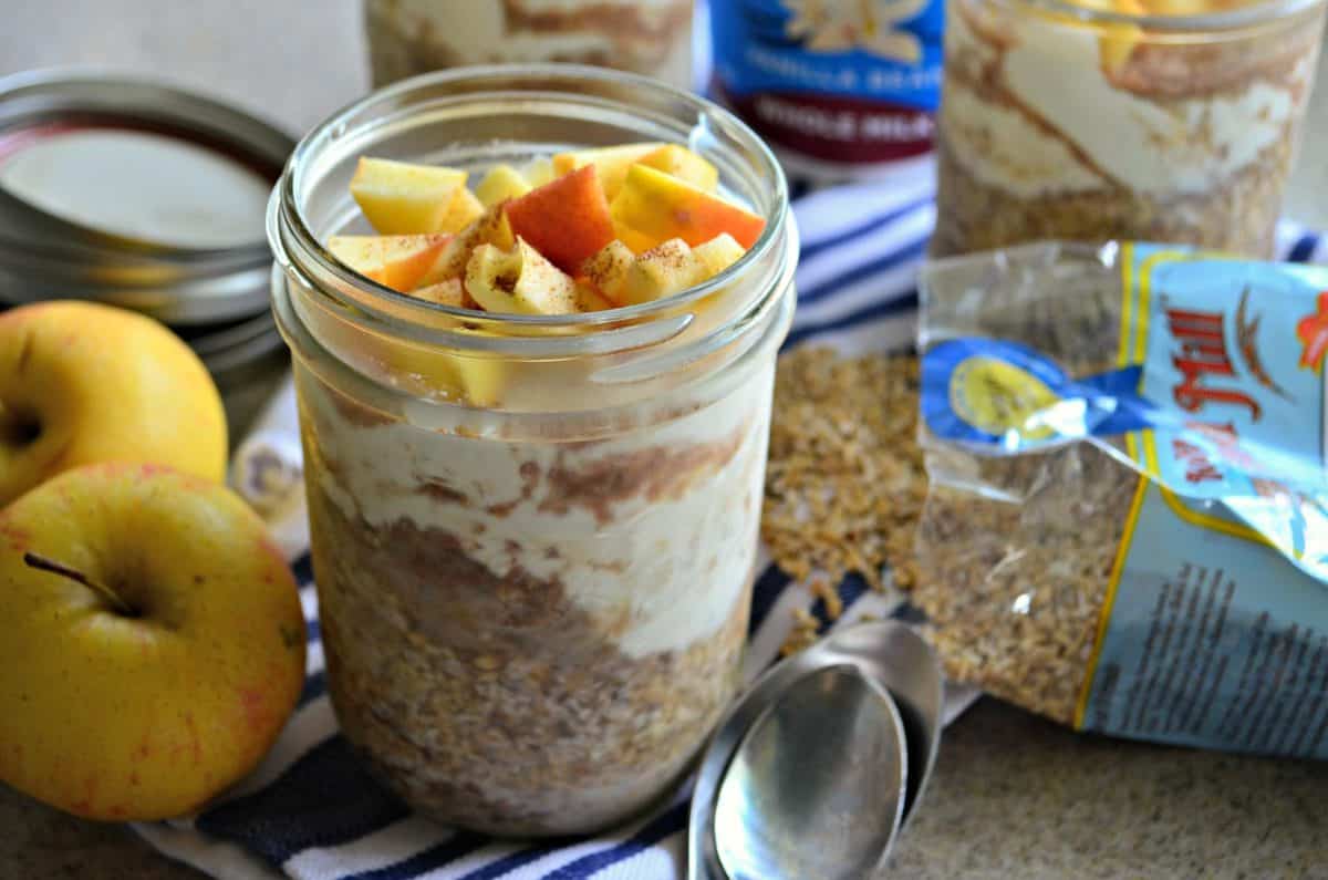 glass filled with yogurt, oats, and topped with apple slices and cinnamon on cloth next to spoon and apples.