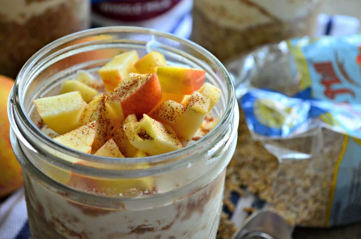 closeup side view glass filled with yogurt, oats, and topped with apple slices and cinnamon on cloth next to spoon and apples.