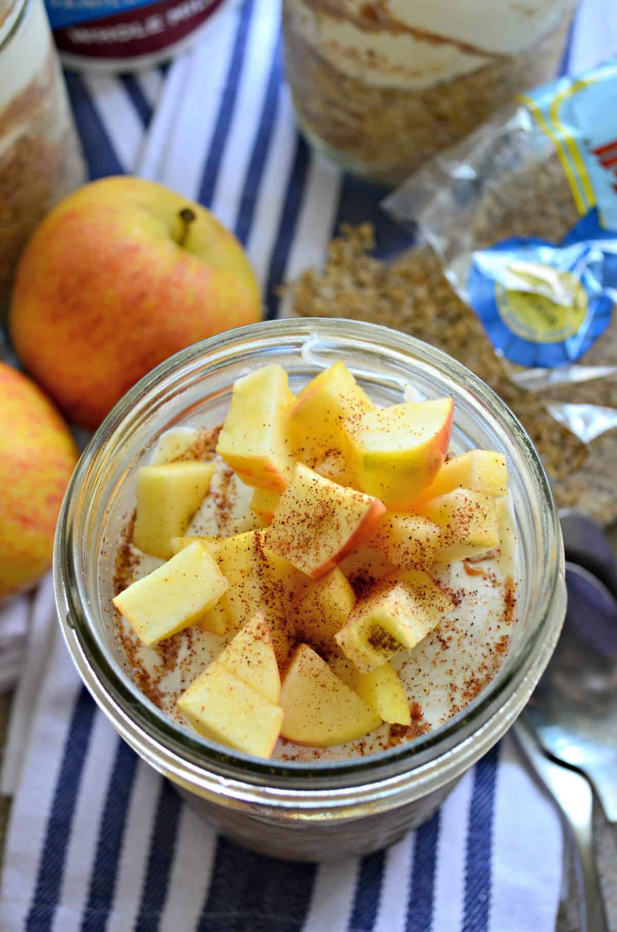 closeup top view glass filled with yogurt, oats, and topped with apple slices and cinnamon on cloth next to spoon and apples.