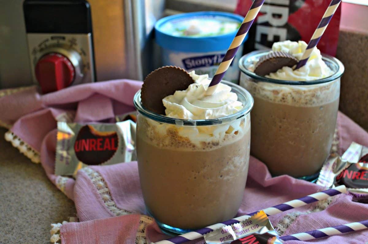 two glasses filled with brown milkshakes topped with whipped cream and peanut butter cups on pink tablecloth.