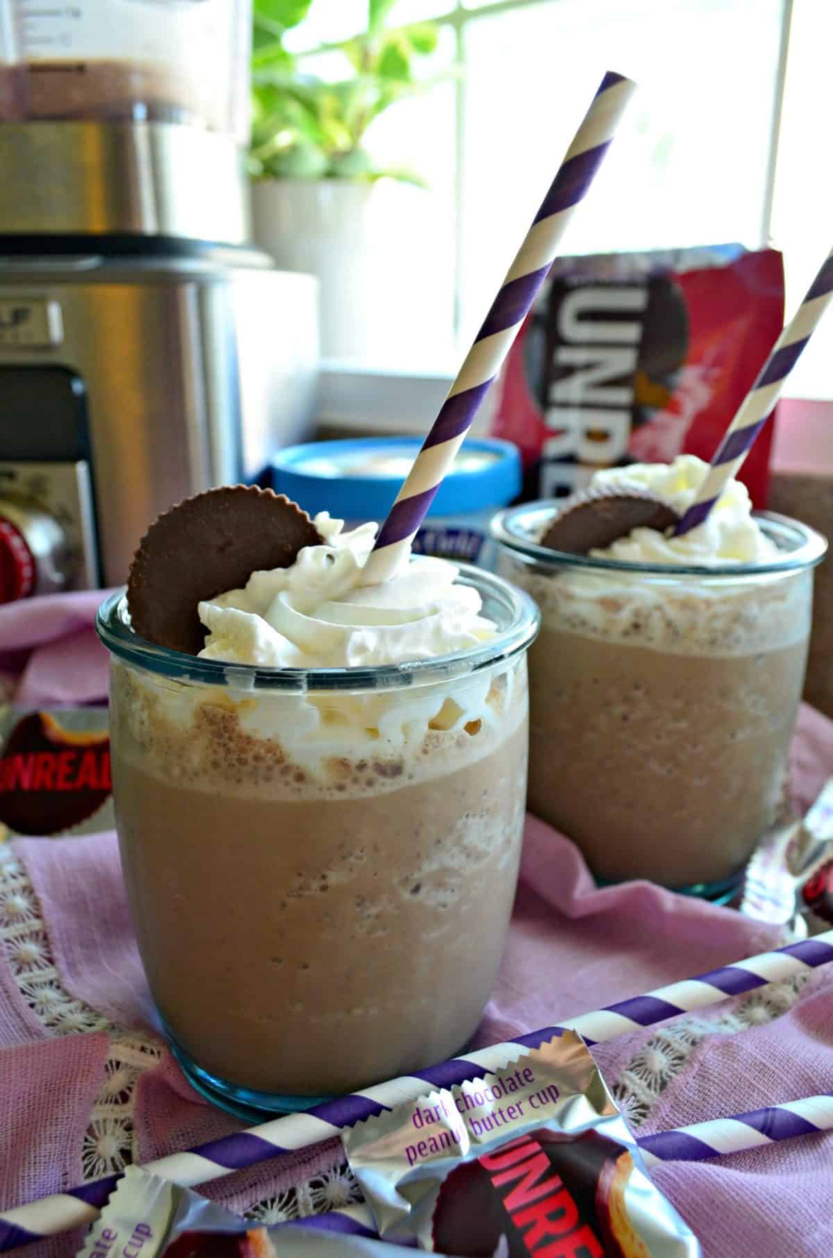 two glasses filled with brown milkshake topped with whipped cream and peanut butter cups on pink tablecloth.