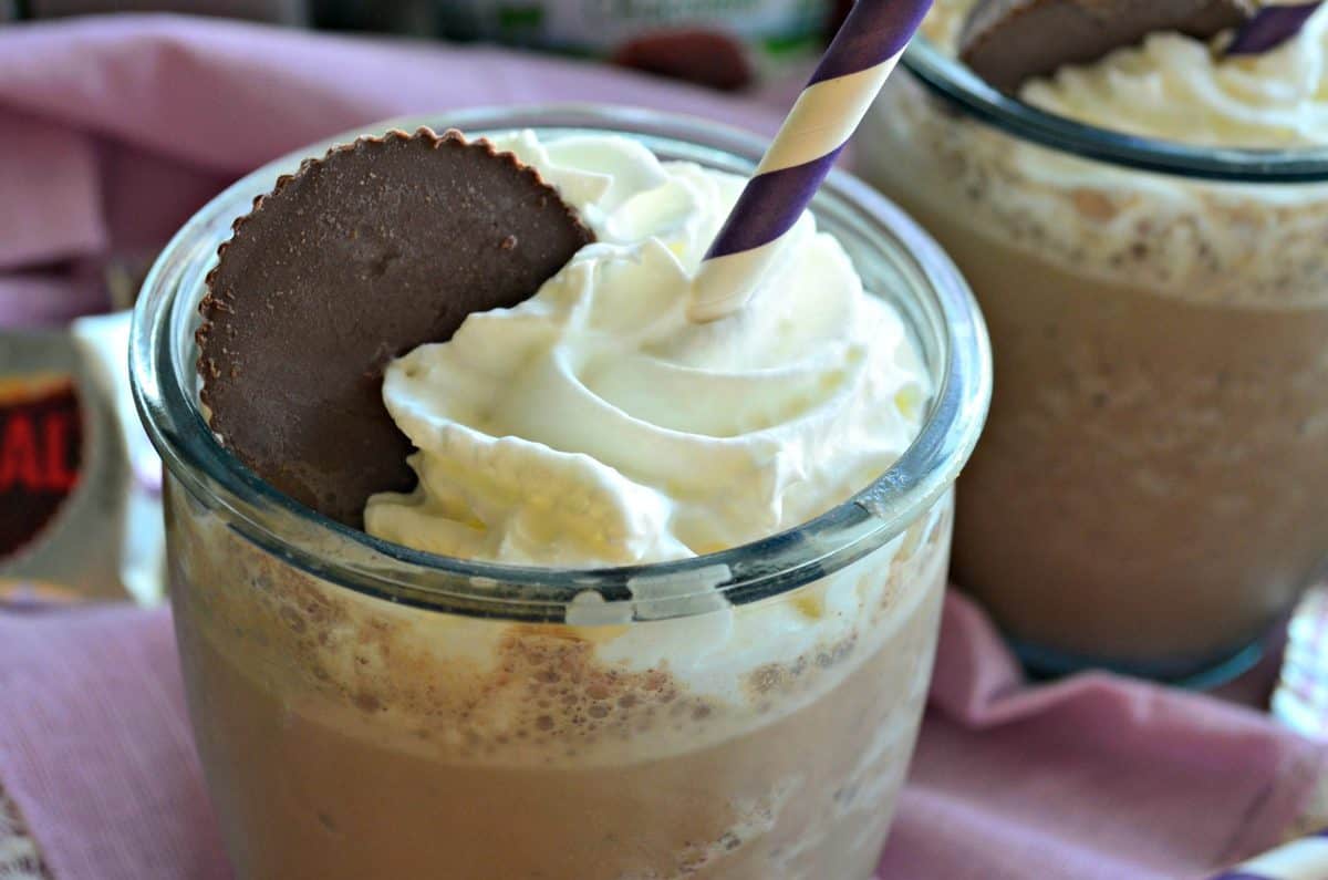 closeup glass filled with brown milkshakes topped with whipped cream and peanut butter cup on pink tablecloth.