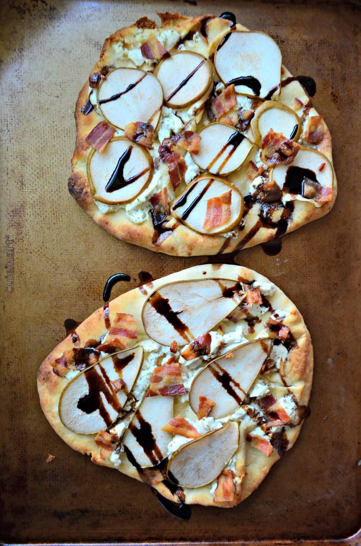 2 Goat Cheese, Bacon, and Pear Naan Flatbreads drizzled with brown sauce on light brown surface.