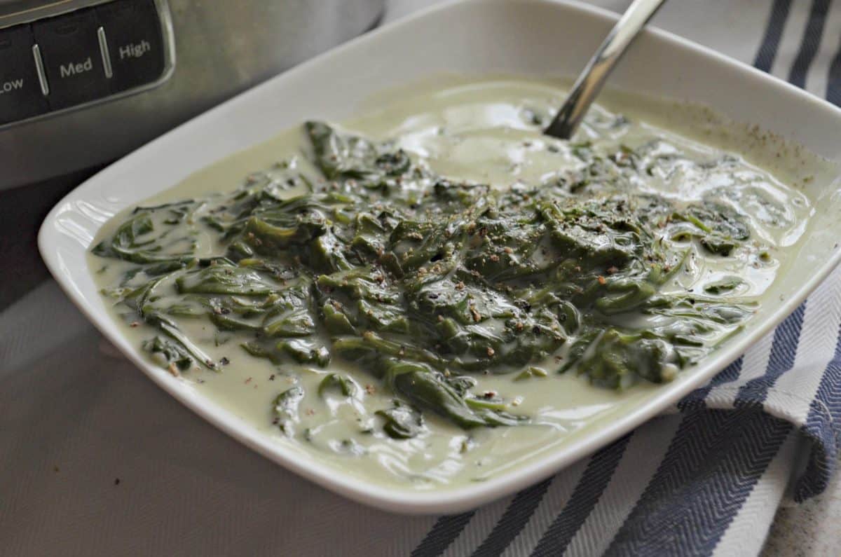 white bowl of Creamed Spinach with spoon in front of slow cooker.