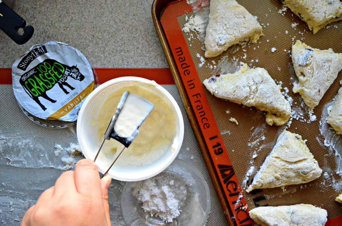 top view of unbaked sliced Cranberry Vanilla Yogurt Scones on baking sheet next to scoop of yogurt on counter.