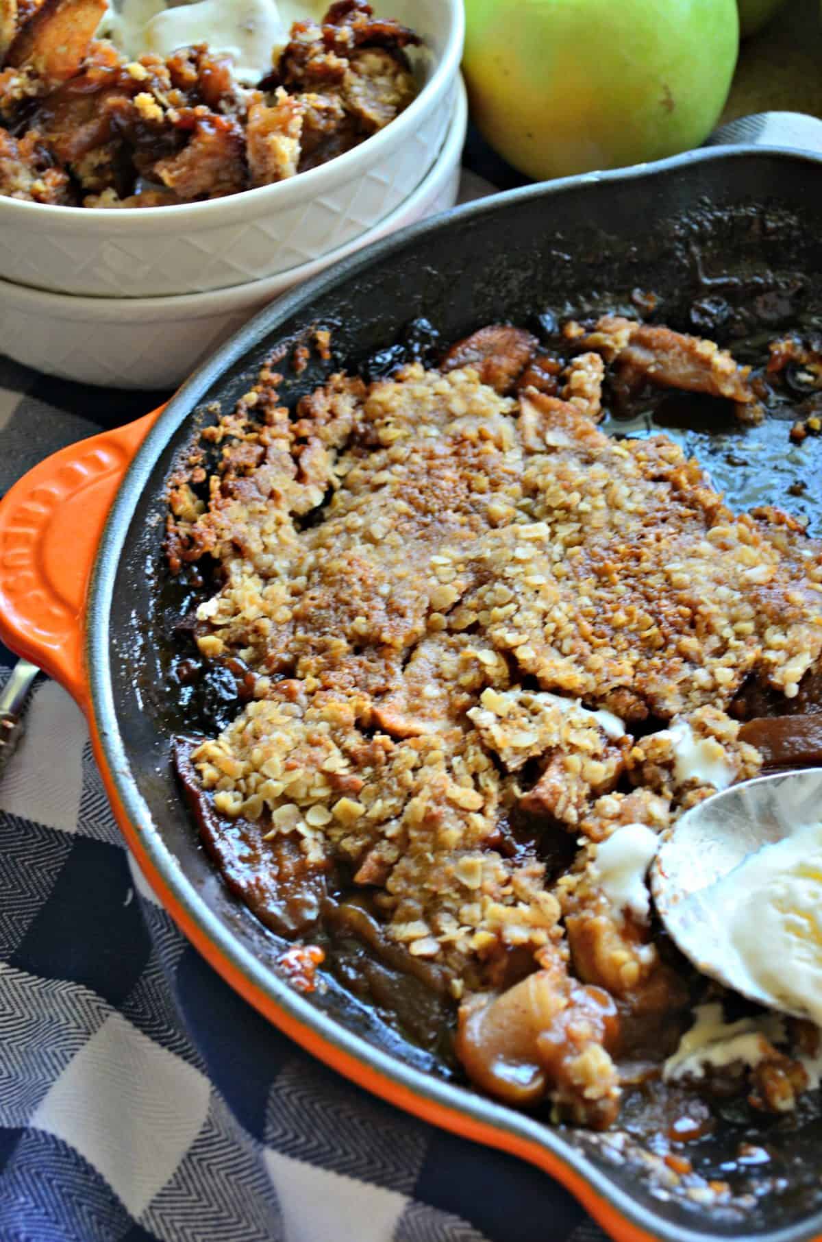 Top view of Apple Crisp in skillet with half spoonful of ice cream on checkered tablecloth. 