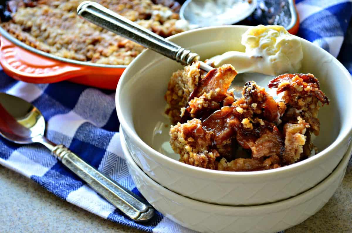 small white bowl of Apple Crisp with vanilla ice cream and spoon on checkered tablecloth.