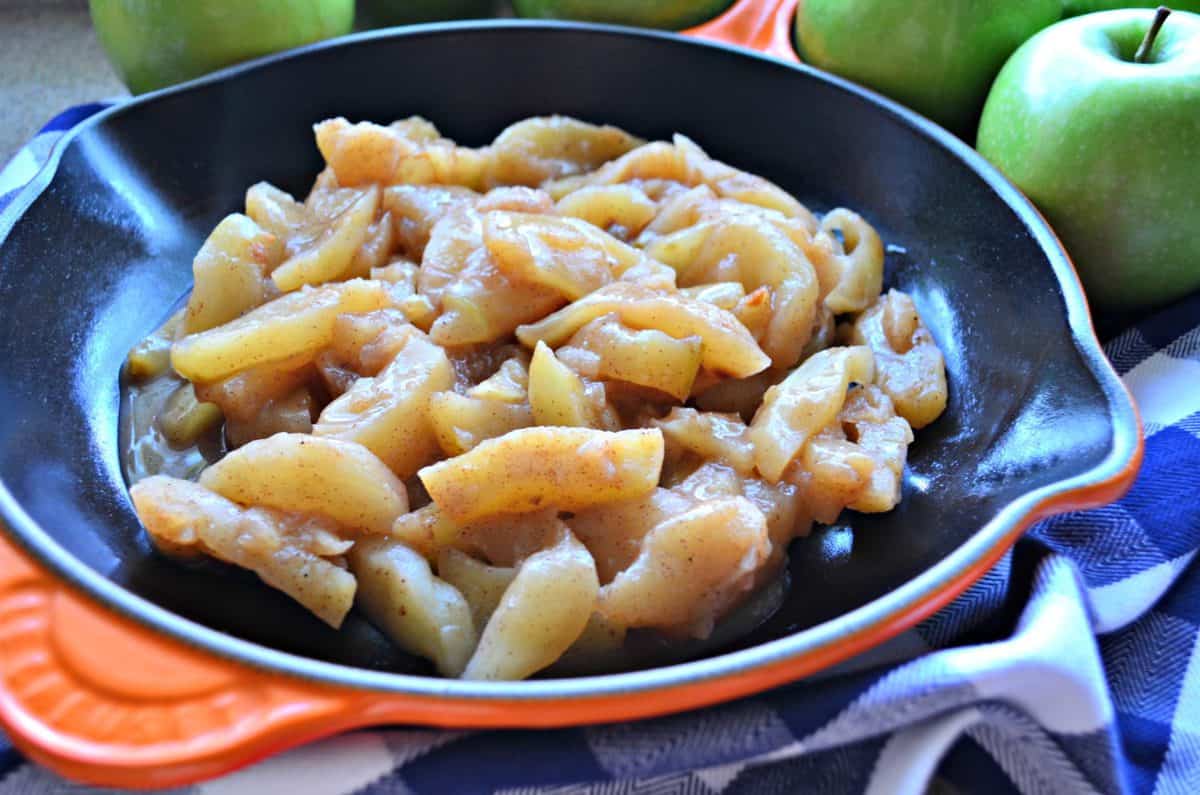 Orange cast iron skillet with cooked cinnamon apples with a blue and white checkered cloth next to it.