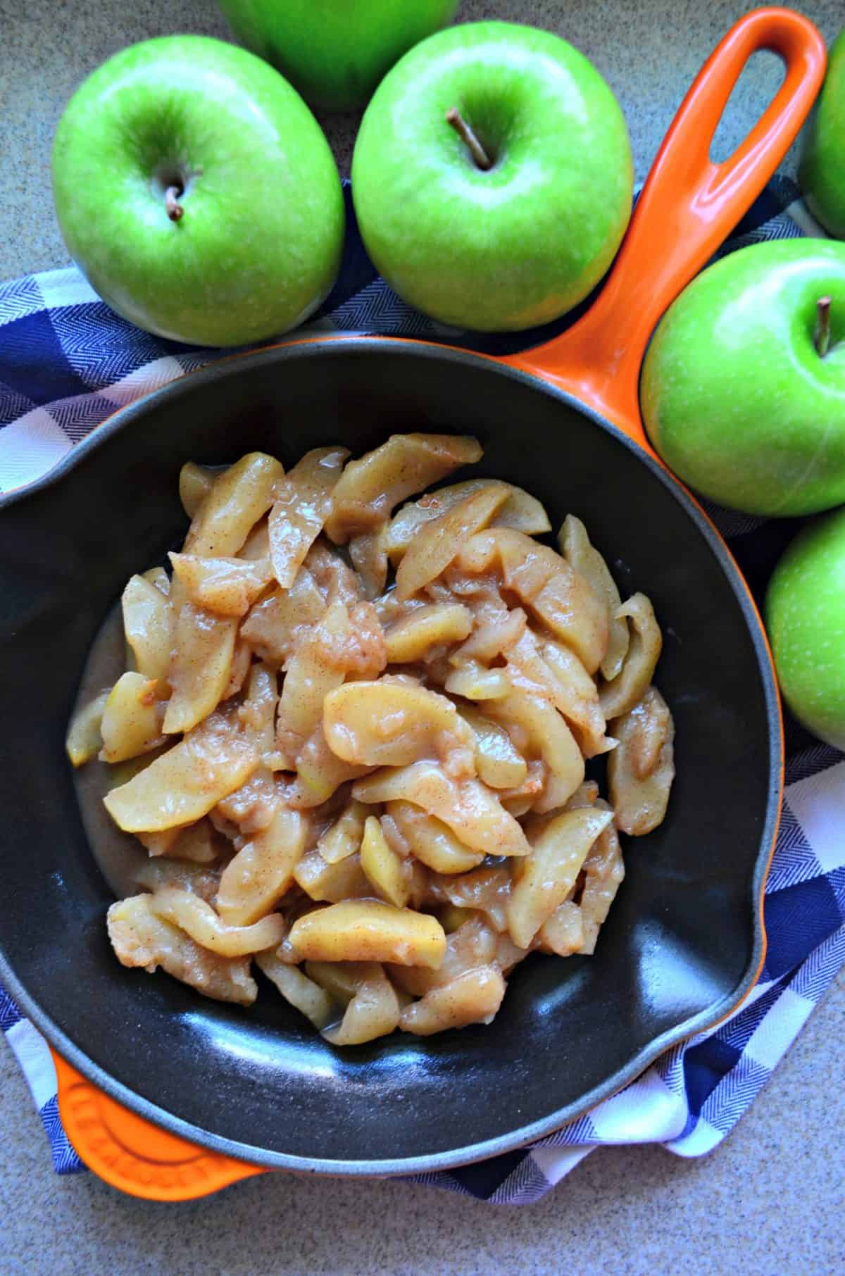 Top view of an orange cast iron skillet filled with cooked cinnamon apples and fresh granny smith apples next to the skillet.