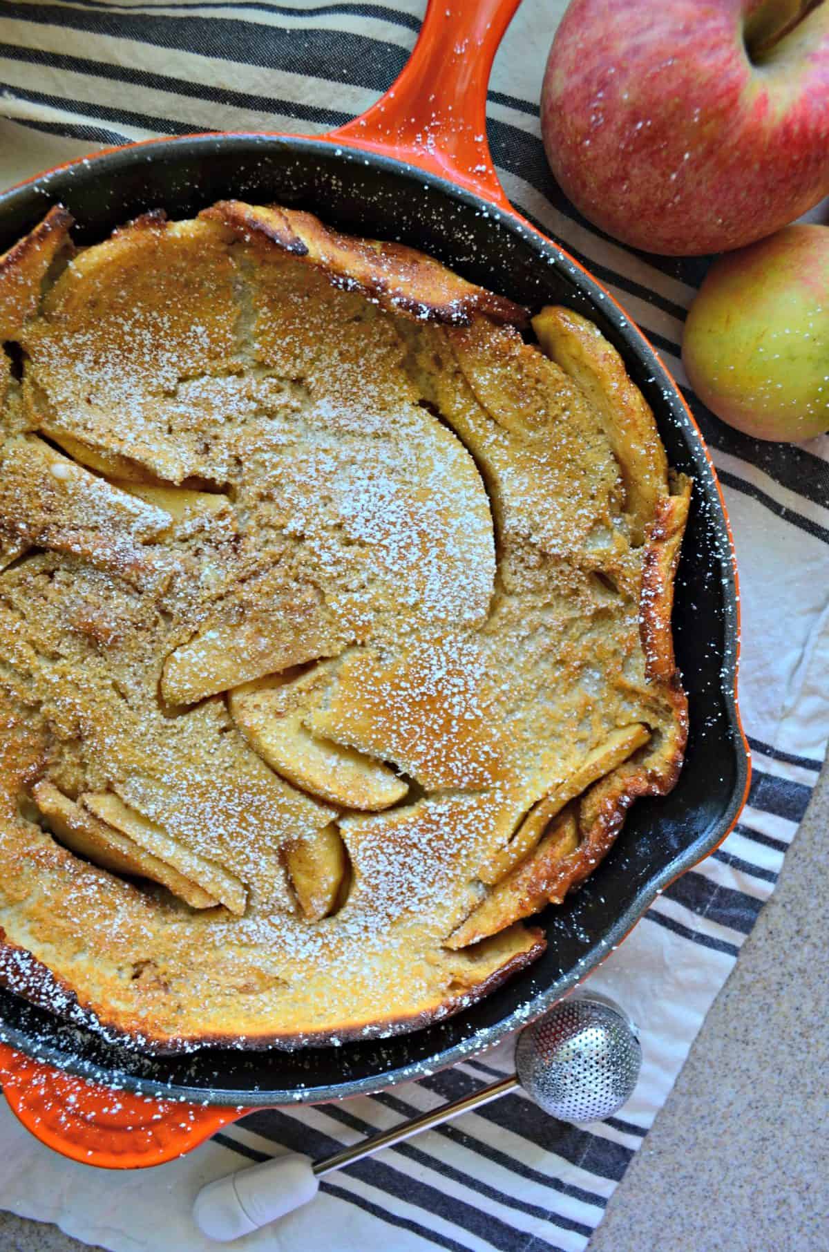 top view of pancake-like pastry with apple slices baked in skillet, topped with powdered sugar.