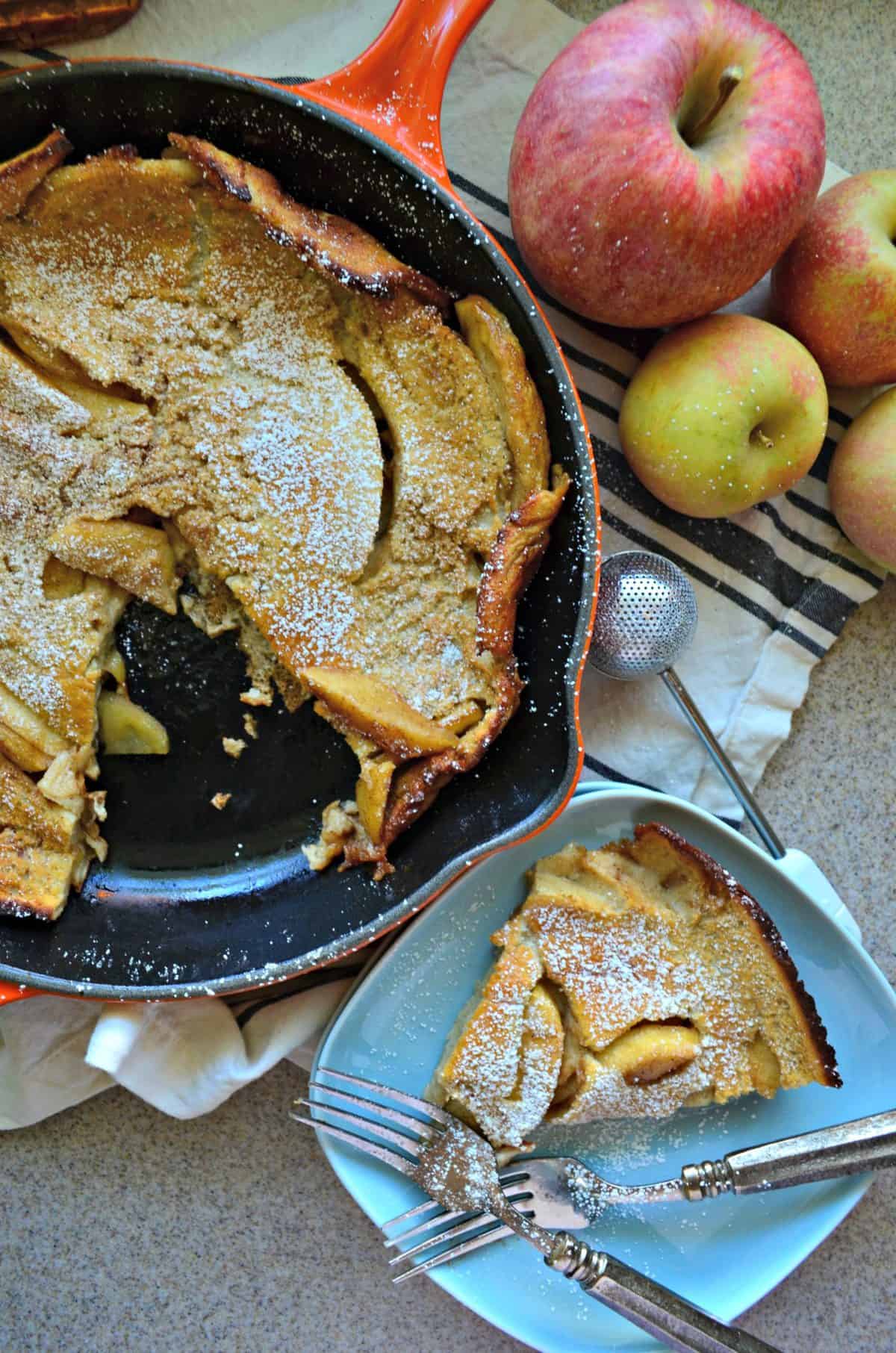 top view of pancake-like pastry with apple slices baked in skillet, topped with powdered sugar next to plated slice.