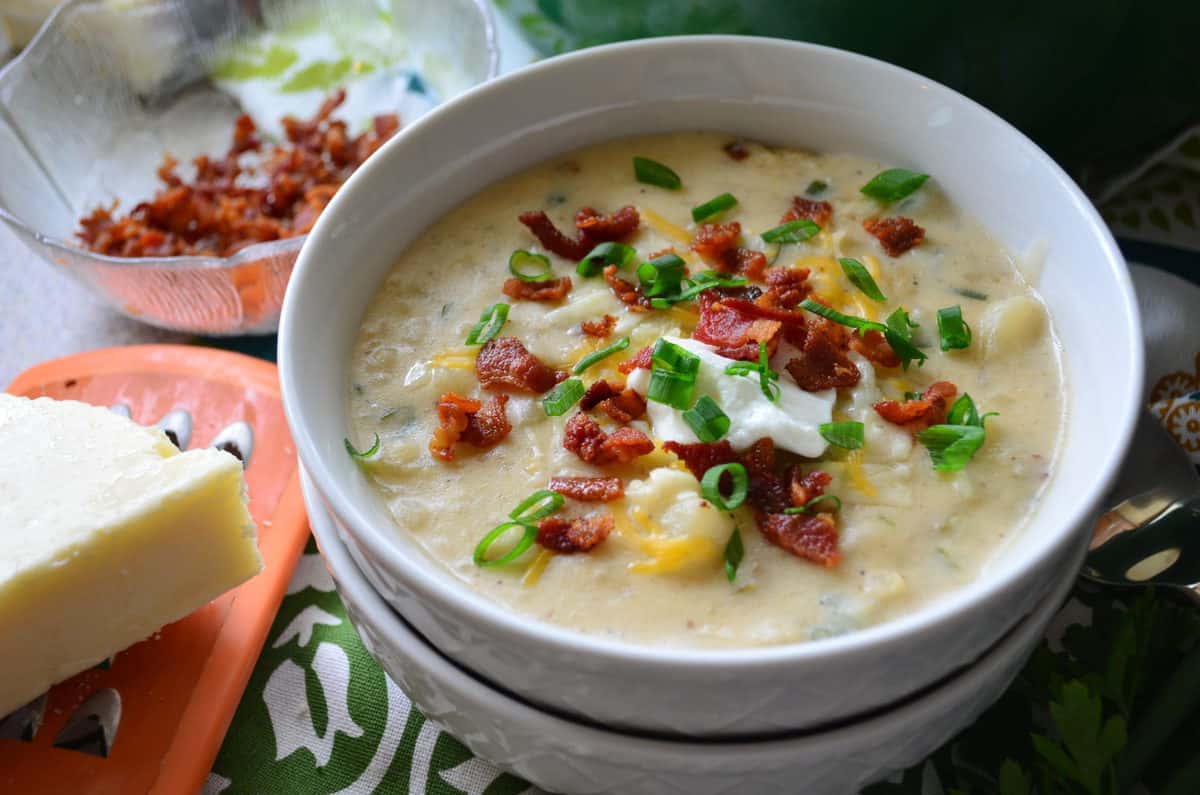 potato soup in white bowl topped with chives, bacon bits, cheese, and sour cream next to cheese and grater.