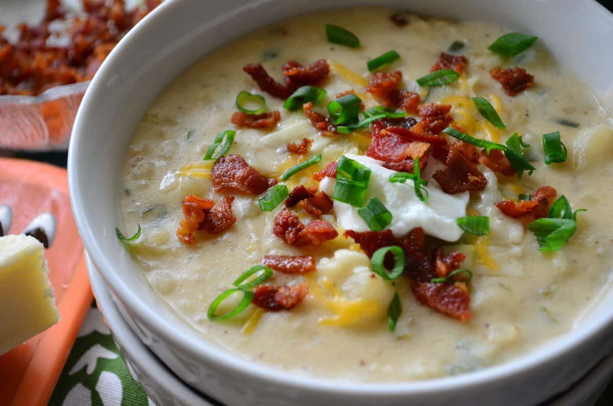 closeup of potato soup in white bowl topped with chives, bacon bits, cheese, and sour cream.