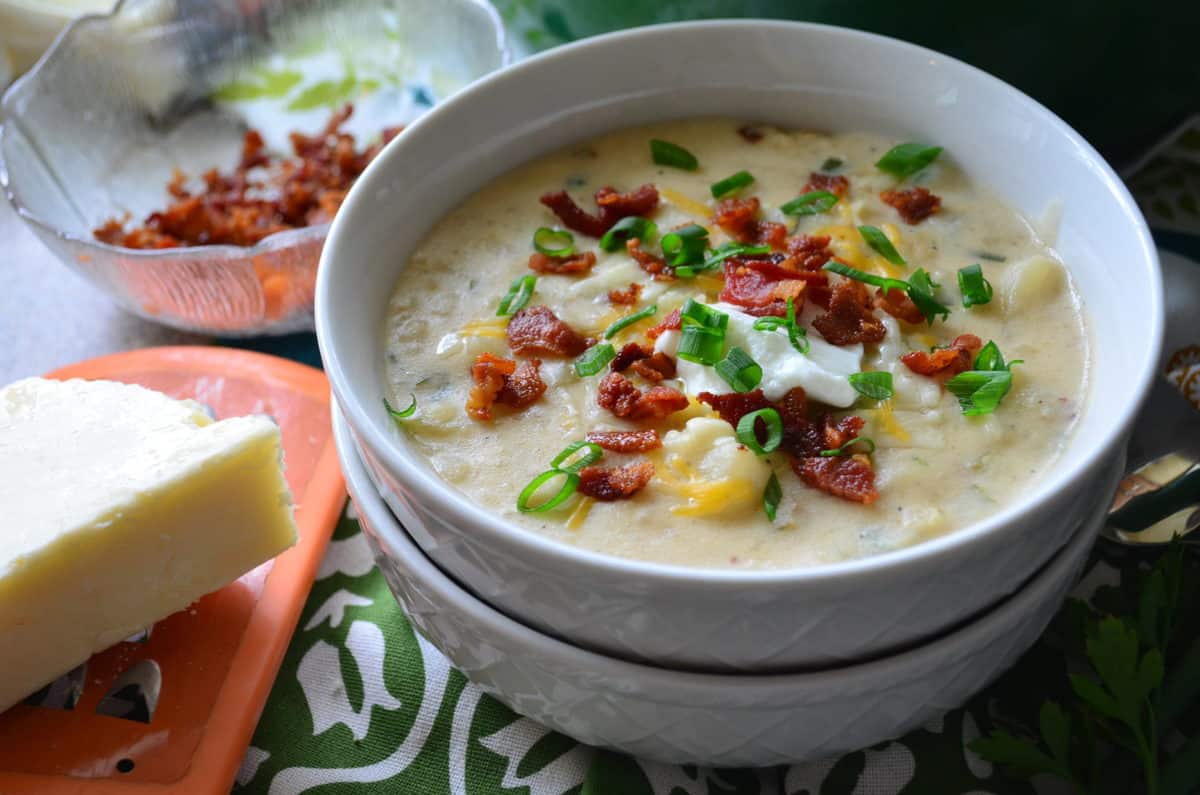 top view of potato soup in white bowl topped with chives, bacon bits, cheese, and sour cream on tablecloth.