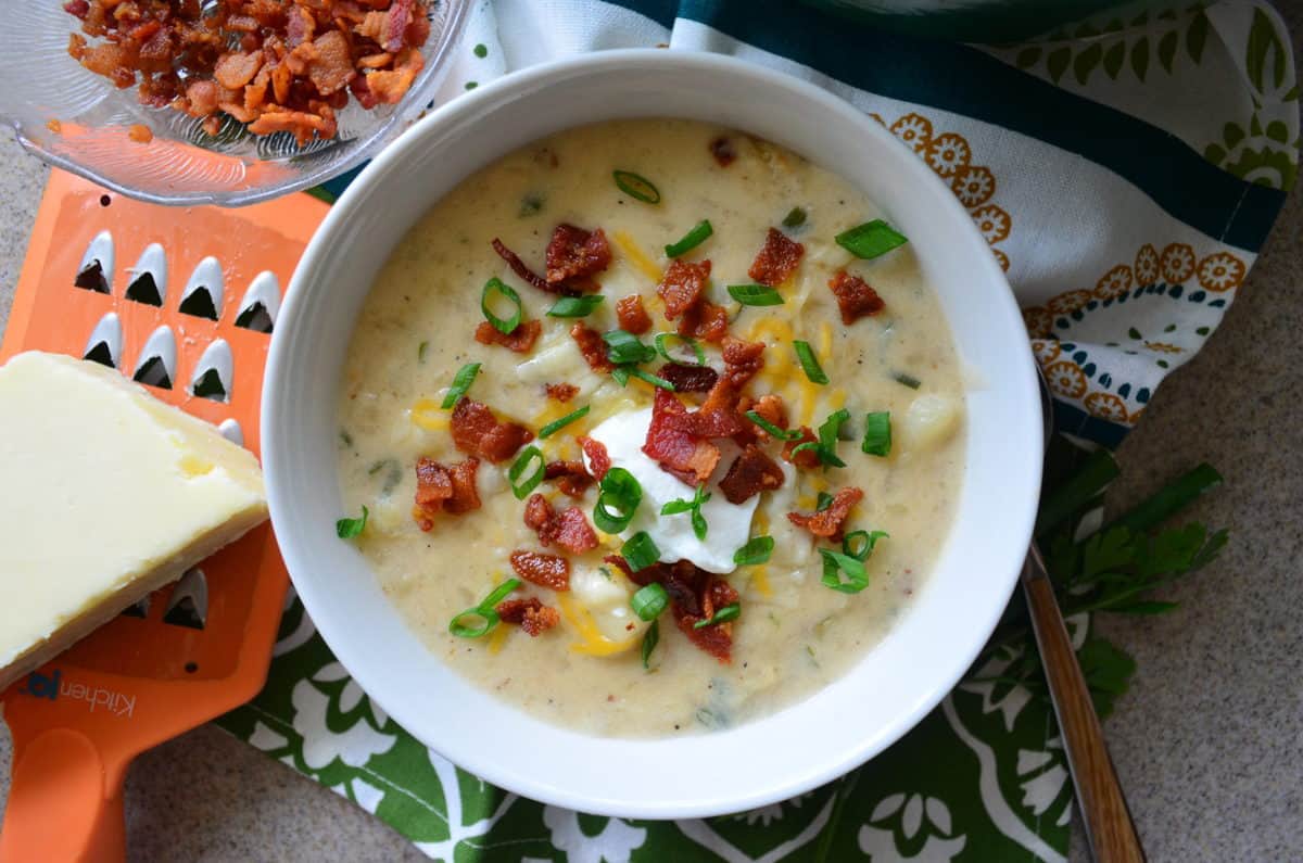 top view of potato soup in white bowl topped with chives, bacon bits, cheese, and sour cream next to cheese and grater.