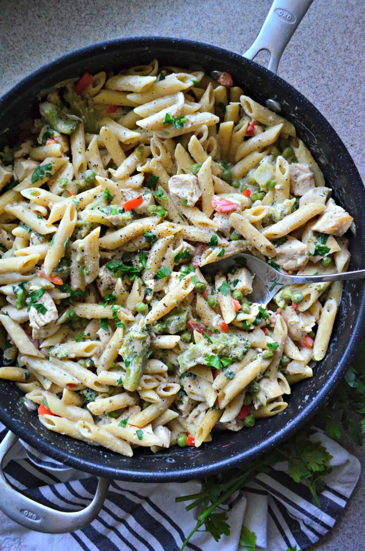 top view skillet of penne with peas, herbs and spices, bell peppers, white meat, and broccoli with spoon in it.