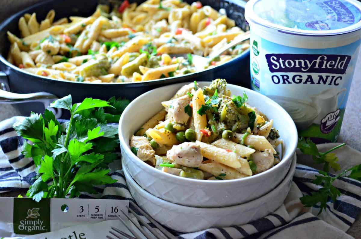 bowl and skillet of penne with peas, herbs and spices, bell peppers, and broccoli near yogurt and parsley.