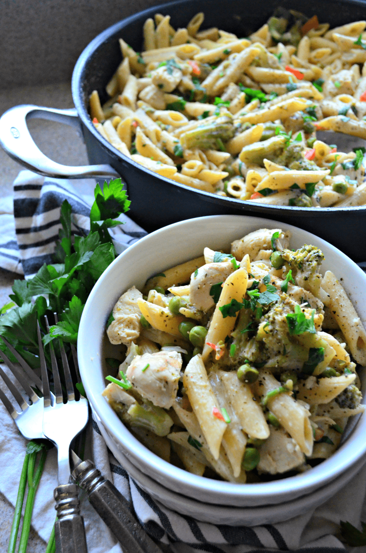 bowl and skillet of penne with peas, herbs and spices, white meat, bell peppers, and broccoli next to fresh parsley.